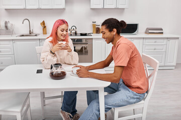 good looking cheerful diverse couple sitting at table with coffee and looking at each other