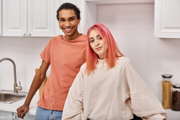 good looking cheerful diverse couple posing together and smiling at camera in kitchen at home