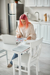 jolly young woman in casual attire with pink hair working with laptop and paperwork with images