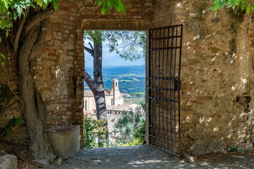 View through the opening of a door in the fortified wall in Parco della Rocca of San Gimignano, Italy with view on Chiesa di Sant Agostino and typical Tuscan landscape - obrazy, fototapety, plakaty