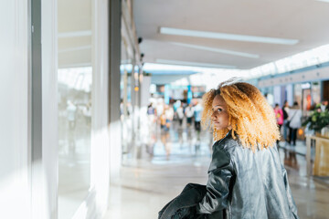 A woman with curly hair is walking through a mall. She is wearing a black jacket and she is looking at windows. The mall is crowded with people, and there are several potted plants and a bench in the 