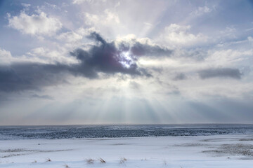 Panoramic winter landscape in Iceland with white and black clouds over snowy and icy flat lava rock landscape with bright sun behind black cloud with strong sunrays