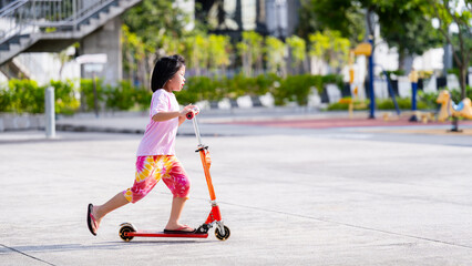 Side view of Asian Child Girl having fun Riding a red scooter at a playground, warm sunlight, in summer or spring time, copy space, leisure activities, exercise kid, Children aged six years old. © Kanthita
