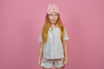 Obraz na płótnie Canvas Funny redhead teen girl makes faces wearing sleeping mask on pink background.