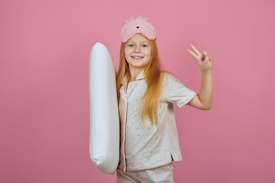 Smiling redhead teen girl holding pillow and wearing sleeping mask on pink background.