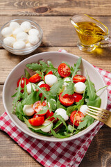 Fresh summer vegetable salad with mozzarella cheese, green arugula leaves and cherry tomatoes on wooden table background. Healthy food, diet concept. Flat lay, top view.