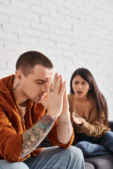 young tattooed man sitting with closed eyes near angry asian man quarrelling at home, divorce