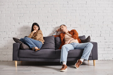 young self-confident man talking to upset asian wife on couch in living room, divorce concept - obrazy, fototapety, plakaty