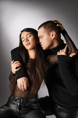 Fototapeta premium trendy interracial couple in black casual attire embracing with closed eyes on grey backdrop