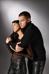 passionate man in black attire embracing alluring asian woman on grey backdrop, love and fashion