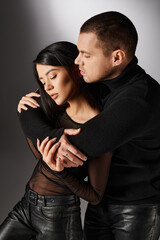 handsome man in black outfit embracing alluring asian woman on grey backdrop, love and fashion
