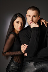 fashionable expressive interracial couple in black clothes looking at camera on grey backdrop