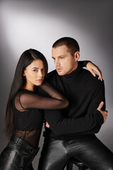 affectionate asian woman embracing trendy man in black casual attire sitting on chair on grey