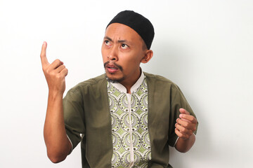 An Indonesian Muslim man in a koko shirt and peci looks confused, pointing his finger upwards while...