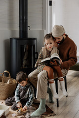 A father and his daughter are reading a book together, while the other daughter is sitting on the floor at his feet and playing with toys