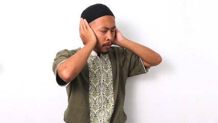 Indonesian Muslim man in koko and peci raises his hands to his ears in the traditional gesture of...