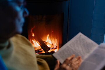 Senior woman, wrapped in warm knitted plaid, relaxing at home, sitting in chair by fireplace and...