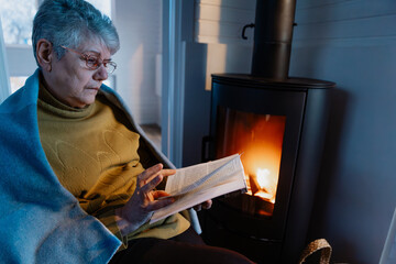 Senior woman, wrapped in warm knitted plaid, relaxing at home, sitting in chair by fireplace and...