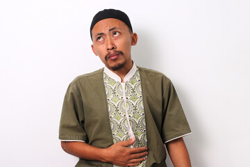 An Indonesian Muslim man in koko and peci holds his stomach, indicating hunger while he awaits...
