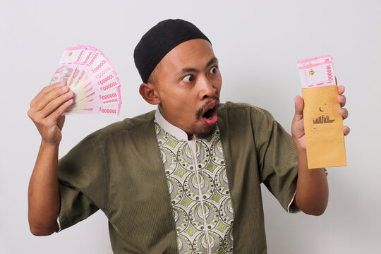 A surprised Indonesian Muslim man looks at a brown envelope filled with Indonesian Rupiah banknotes, representing his Angpao Lebaran or Eid al-Fitr money gift. Isolated on a white background