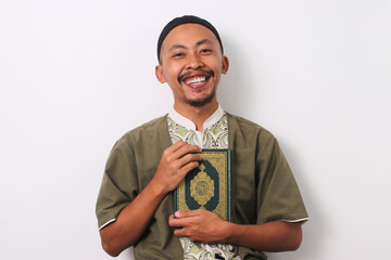 A joyous Indonesian Muslim man in koko and peci holds the Holy Quran to his chest, smiling warmly at the camera. Isolated on a white background