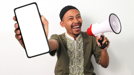 Excited Indonesian Muslim man in koko and peci shows a blank phone screen and holds a megaphone, promoting a new app or Ramadan sale. Isolated on a White background - obrazy, fototapety, plakaty