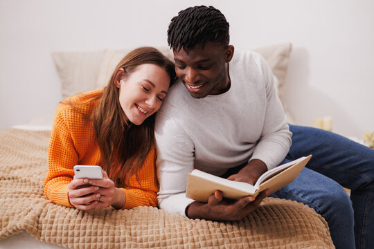 Joyful multiethnic couple with book and smartphone relaxing on bed at home