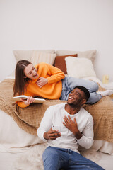 Cheerful multiethnic couple with book and smartphone relaxing in bedroom 