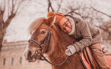 a boy sitting in a saddle lies on the neck of a pony and smiles looking to the side
