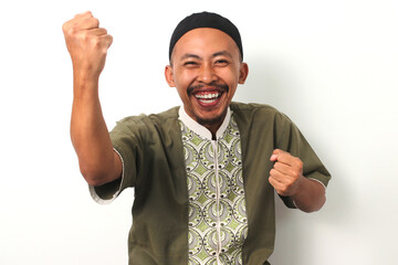 An Indonesian Muslim man in koko and peci raises his fist in a gesture of joy and celebration,...