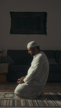 Side vertical wide footage of bearded muslim man in white sitting alone on floor in spacious room and praying takbirs while reciting Koran in silence at home