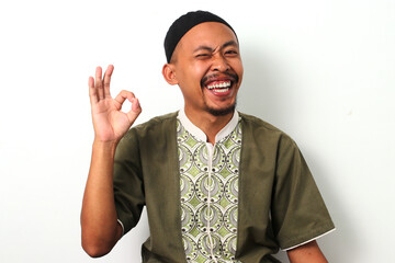 Happy Indonesian Muslim man in koko and peci gives a playful wink and an OK gesture, expressing...