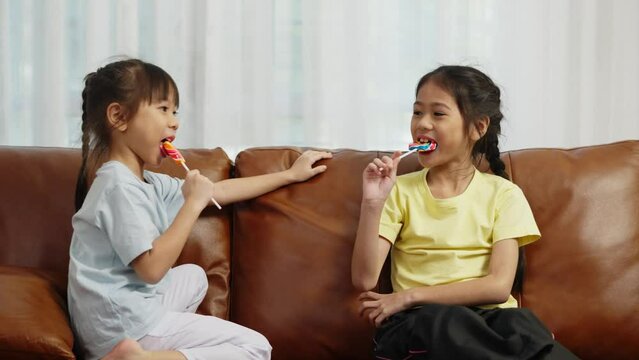 Footage handheld medium shot, Two cute girls sitting on sofa and enjoy to play and eating big lollipop together, Asian sibling girls talking with fun in living room at home