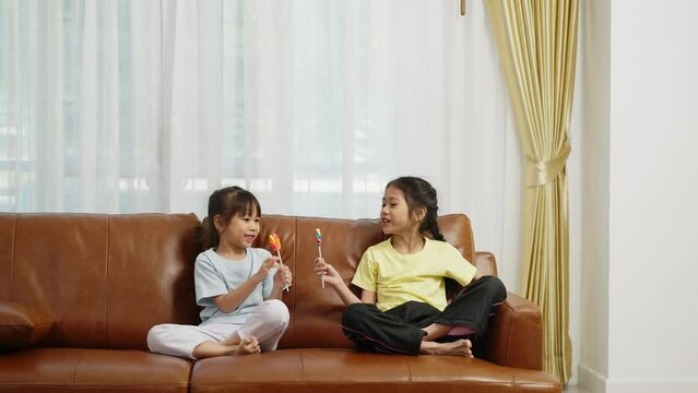 Footage handheld wide shot, Two cute girls sitting on sofa and enjoy to play and eating big lollipop together, Asian sibling girls talking and jump with fun in living room at home