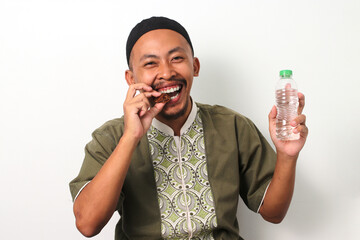 A happy Indonesian Muslim man in a koko shirt and peci holds dates and mineral water, ready to...