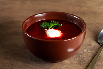 brown Bowl of tasty borscht with sour cream and greens with spoon on wooden background