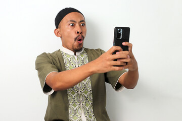 Shocked Indonesian Muslim man in koko shirt and peci stares at his phone in disbelief. Isolated on a white background