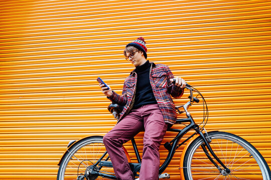Young hipster man using mobile phone while sitting on cruiser bicycle near orange wall. Student using 5g network,rests after riding. Eco friendly transport for carbon footprint. Sustainable lifestyle