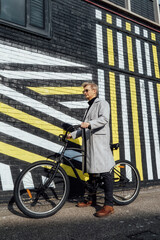 Fototapeta na wymiar Stylish male in checked coat and sunglasses standing with retro bicycle by painted urban wall in sunny day. Neutral carbon footprint transportation. Green eco friendly mobility sustainable transport