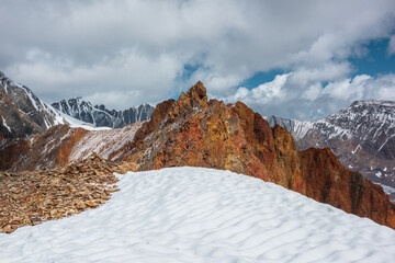 Snowfield on stony mountain ridge and vivid sharp pinnacle in sunlight. Shiny pointy peak of gold color. Colorful red rocky peaked top. Freshly fallen snow in high mountains. Sunny cloudy alpine view.
