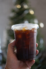 Man hand holding a black coffee in a clear glass whit Christmas tree behide.
