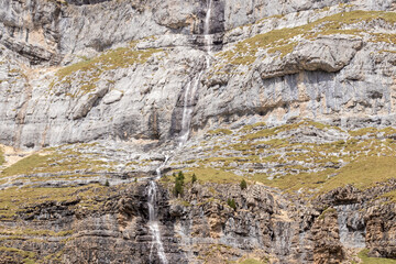 a waterfall cascading down a rugged, rocky mountain, surrounded by sparse green vegetation under a cloudy sky.