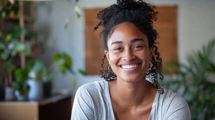 A young biracial woman smiles warmly on video call, seated indoors.