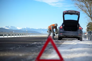 Fotobehang the driver of a motor vehicle had a car breakdown while driving on the road in winter, put a warning triangle on the road and tries to change a wheel on the car. © juraj