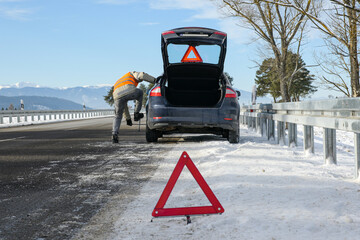 the driver of a motor vehicle had a car breakdown while driving on the road in winter, put a warning triangle on the road and tries to change a wheel on the car.