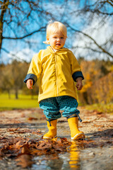 Sun always shines after the rain. Small bond infant boy wearing yellow rubber boots and yellow waterproof raincoat walking in puddles in city park on sunny rainy day - 748795043