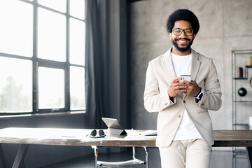 Young businessman in a modern workspace smiles and holds a smartphone, signifying the positive...