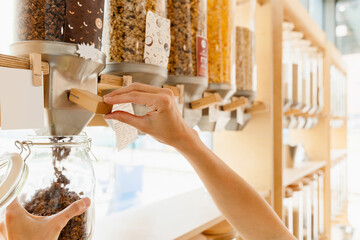 Female customer filling dry cereals in glass jar from dispenser at zero waste shop. Sustainable shopping in modern plastic free grocery store.