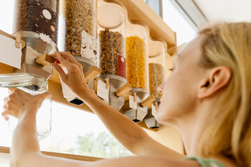 Female customer filling dry cereals in glass jar from dispenser at zero waste shop. Sustainable shopping in modern plastic free grocery store.