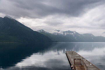 A wooden pier juts into the reflective fjord, drawing the eye towards the overcast Norwegian...